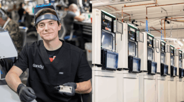 Dandy dental lab technician holding a crown beside a row of dental milling machines in the lab