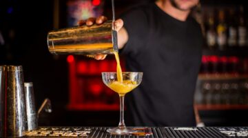 A bartender pouring a yellow cocktail from a metal shaker into a coupe glass at a dimly lit bar.