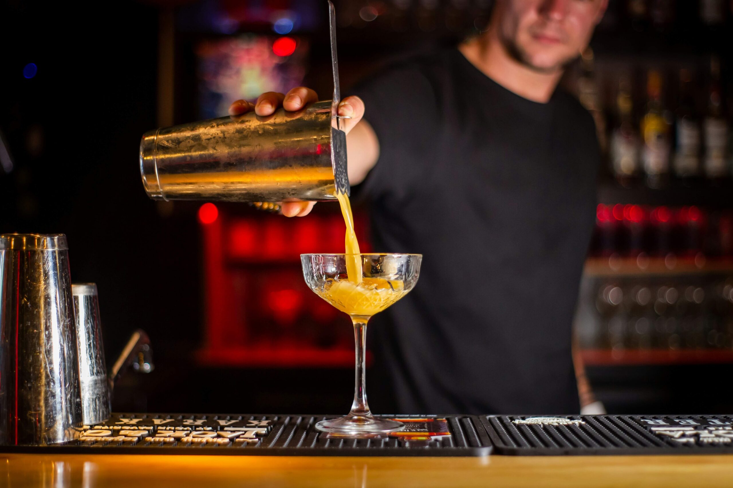 A bartender pouring a yellow cocktail from a metal shaker into a coupe glass at a dimly lit bar.